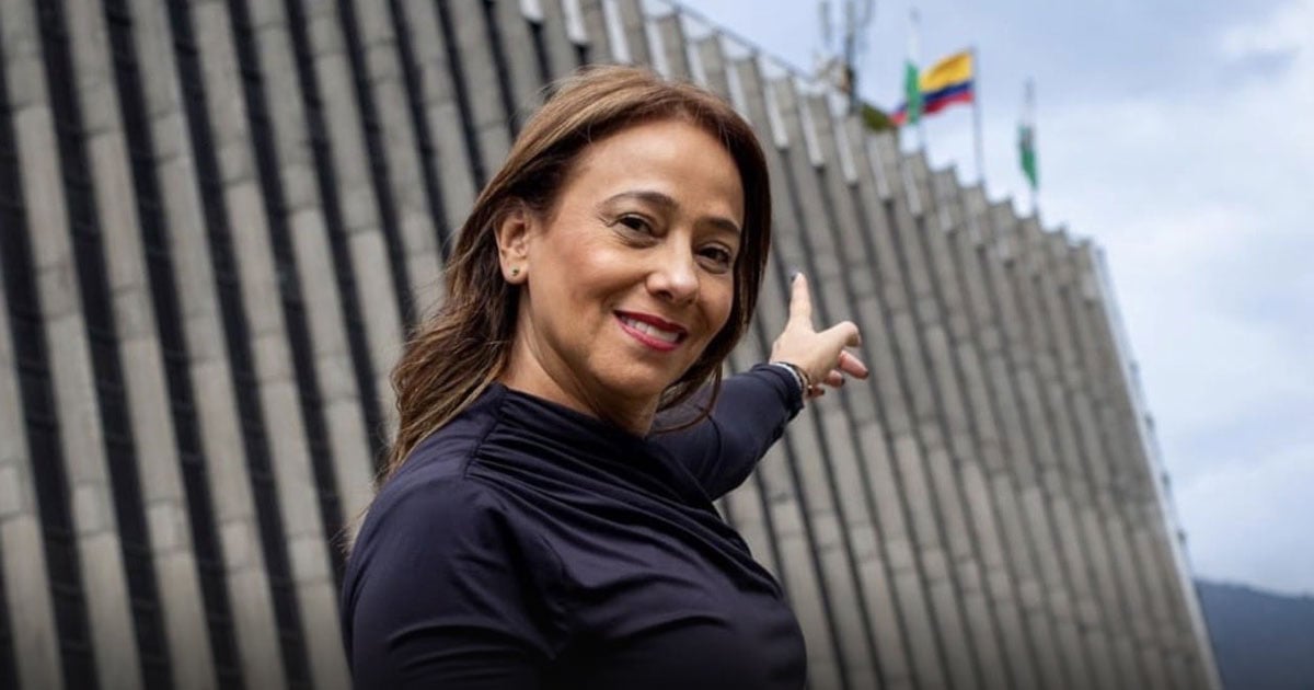 Smiling woman in a dark top gestures toward a slanted, striped wall with several flags on top. - El salto de Claudia Carrasquilla: de concejal de Medellín a viceministra de Defensa
