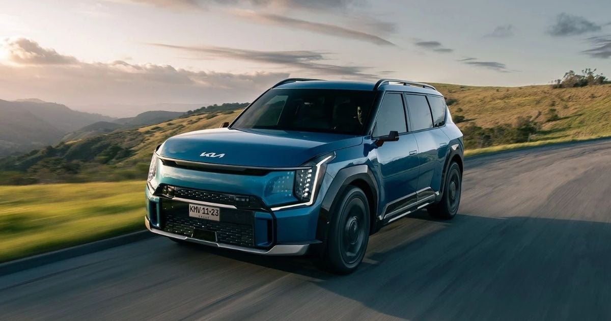 Blue SUV (front view) driving along a winding rural road at sunset with rolling hills in the background. - La camioneta eléctrica de Kia que parece sacada del futuro: seis pasajeros, potente y con casi 500 km de autonomía