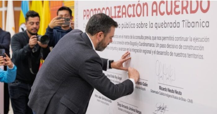 Vía libre para la construcción del puente Tibanica en Bosa, beneficiará a 250 mil personas de Ciudad Verde Man in a suit signs a large wall-mounted agreement as photographers film the moment, in front of a banner reading 'PROTOCOLIZACIÓN ACUERDO'. - Vía libre para la construcción del puente Tibanica en Bosa, beneficiará a 250 mil personas de Ciudad Verde