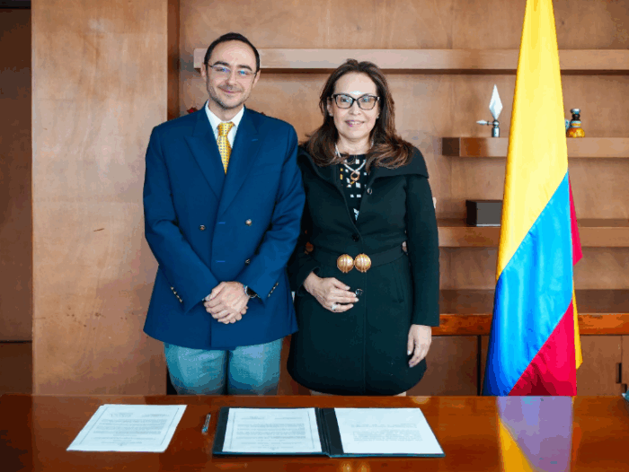 Con Viviane Morales al frente, los cristianos aterrizan con cargos de poder en el Ministerio de Educación Two professionals stand behind a table with signed documents, a Colombian flag visible on the right. - Con Viviane Morales al frente, los cristianos aterrizan con cargos de poder en el Ministerio de Educación