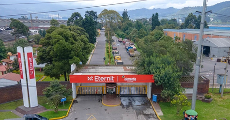 Con Eternit para casas en Buenaventura el millonario Carlos Slim se une a la reconstrucción Aerial view of the entrance to an Eternit factory site with a red sign and gate, lined by trees and a road leading into the complex. - Con Eternit para casas en Buenaventura el millonario Carlos Slim se une a la reconstrucción