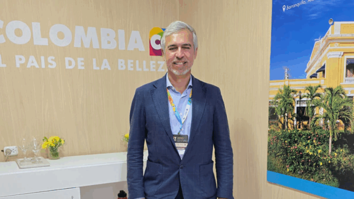 Man in a navy blazer with a conference badge, smiling, standing indoors next to a wall with COLOMBIA lettering and a palm-tree photo backdrop and yellow building. - Quiénes están en los mejores oficinas de Procolombia en el exterior que El Tigre va a acabar
