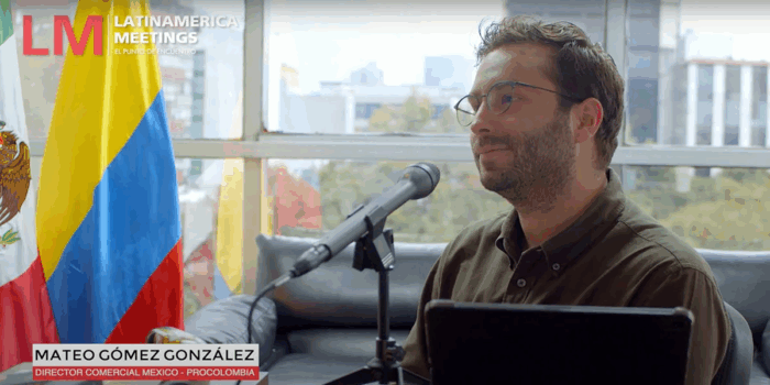 Man with glasses speaks into a microphone during an interview, with the LatinAmerica Meetings logo and a Colombian flag in the background. - Quiénes están en los mejores oficinas de Procolombia en el exterior que El Tigre va a acabar