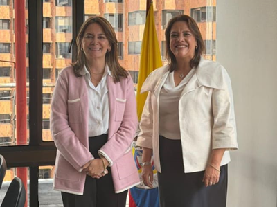 Two professional women posing for a photo indoors, one in a pink blazer and white blouse, the other in a cream blazer; a yellow flag stands between them. - Un equipo formado mayoritariamente por mujeres inicia la tarea de recuperar el sistema de salud
