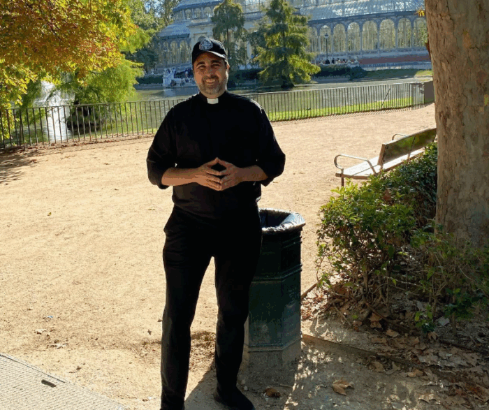 El capellán del Palacio tendrá nuevo rol: será la voz del presidente en 5 universidades públicas Smiling priest in black clerical shirt with white collar stands in a sunny park, hands clasped, with trees and a pond behind him. - El capellán del Palacio tendrá nuevo rol: será la voz del presidente en 5 universidades públicas
