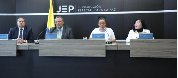 Lo condenaron por matar a Álvaro Gómez, pasó 18 años preso y hoy las Farc dicen que es inocente Cuatro panelistas sentados tras una mesa en una sala de conferencias, con el logo JEP y la bandera de Colombia al fondo. - Lo condenaron por matar a Álvaro Gómez, pasó 18 años preso y hoy las Farc dicen que es inocente