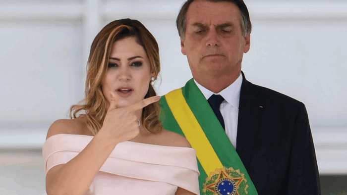 Cómo una atractiva mujer que venía de abajo de quien se enamoró Jair Bolsonaro se volvió el fenómeno político de Brasil Woman in a pale pink off‑the‑shoulder dress points to her face as she speaks, standing beside a man in a suit with a green–yellow sash. - Cómo una atractiva mujer que venía de abajo de quien se enamoró Jair Bolsonaro se volvió el fenómeno político de Brasil