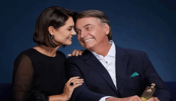 Cómo una atractiva mujer que venía de abajo de quien se enamoró Jair Bolsonaro se volvió el fenómeno político de Brasil Smiling woman and man lean toward each other; she rests a hand on his arm as he holds a perfume bottle and smiles in a dark‑blue backdrop. - Cómo una atractiva mujer que venía de abajo de quien se enamoró Jair Bolsonaro se volvió el fenómeno político de Brasil