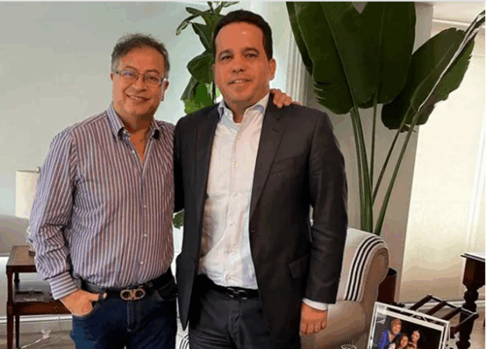 Una movida de la Mintrabajo impidió que el ex senador Carlos Andrés Trujillo pudiera quedarse con Comfenalco Antioquia Two men pose indoors, one in a striped shirt and jeans and the other in a dark suit, with his hand on the other's shoulder, in a bright room with plants and a framed photo nearby. - Una movida de la Mintrabajo impidió que el ex senador Carlos Andrés Trujillo pudiera quedarse con Comfenalco Antioquia