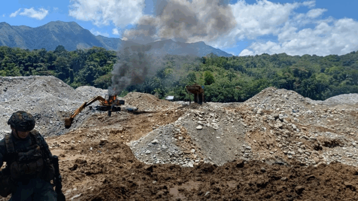 Quarry site with a yellow excavator firing up smoke as rocks pile up, and a person in military gear standing in the foreground. - El oscuro rastro de .600 millones en una empresa de oro manejada por la SAE en el gobierno Petro