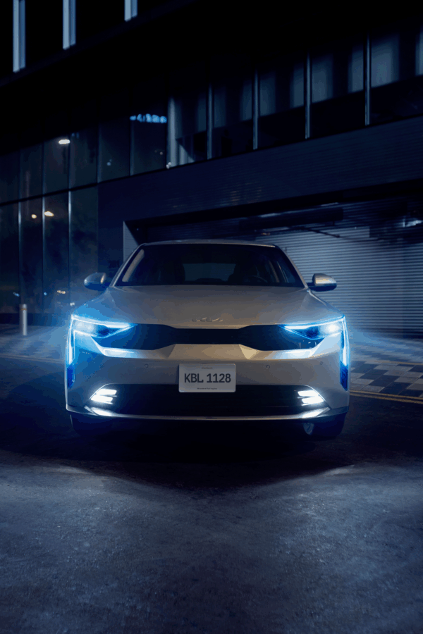 Kia se la juega con un carro que parece más caro de lo que cuesta y quiere conquistar las calles colombianas Front view of a white SUV parked at night in front of a glass-front building with blue-tinted headlights. - Kia se la juega con un carro que parece más caro de lo que cuesta y quiere conquistar las calles colombianas