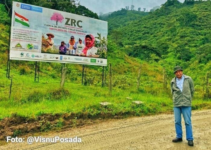 ¿Quiénes son los tres académicos con los que Petro se quería atornillar en la U. Nacional antes de dejar la Presidencia? Man stands on a dirt road beside a large billboard advertising a riverside project, with green hills in the background. - ¿Quiénes son los tres académicos con los que Petro se quería atornillar en la U. Nacional antes de dejar la Presidencia?