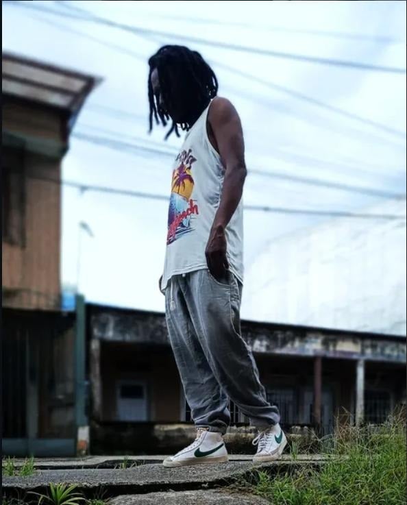 Don Rey, el músico de Quibdó que cambió las armas por instrumentos a cientos de jóvenes del Chocó Young man with dreadlocks wearing a white tropical tank top, gray joggers, and white sneakers with a green swoosh, standing on a curb in an urban area. - Don Rey, el músico de Quibdó que cambió las armas por instrumentos a cientos de jóvenes del Chocó