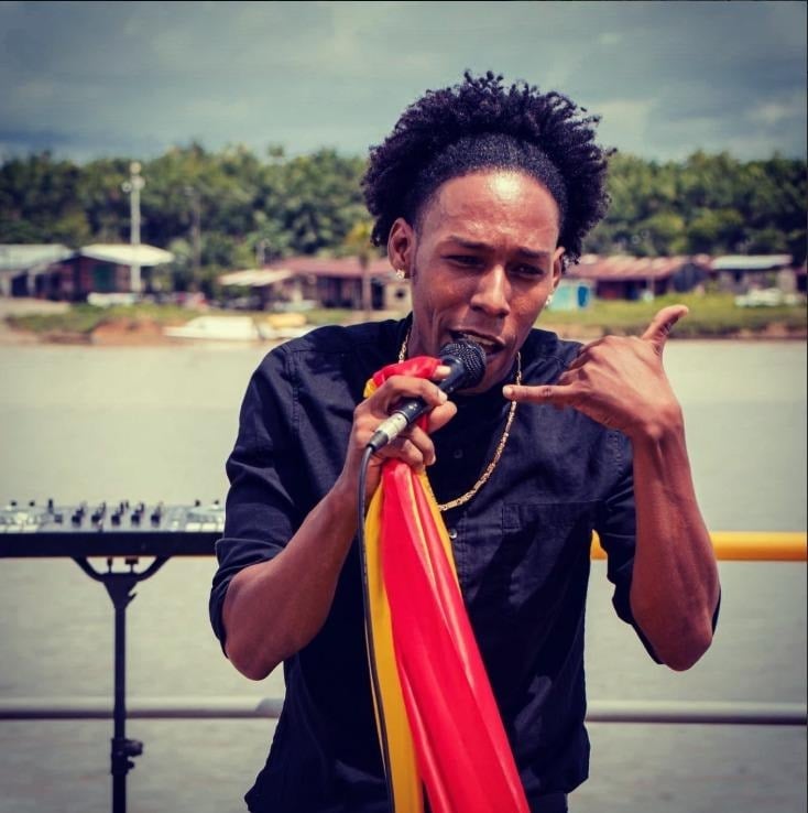 Don Rey, el músico de Quibdó que cambió las armas por instrumentos a cientos de jóvenes del Chocó Male singer performing by a body of water, microphone in hand, wearing a dark shirt and colorful scarf draped over his shoulder. - Don Rey, el músico de Quibdó que cambió las armas por instrumentos a cientos de jóvenes del Chocó