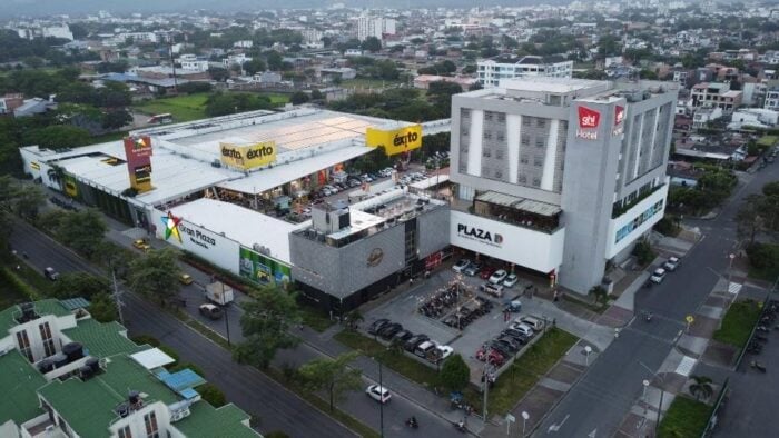 Con un billón de pesos la familia chilena Solari le compró 9 centros a la paisa Argos y Conconcreto Aerial view of a large shopping plaza with multiple storefronts, a parking lot, and a connected tall hotel building with a red 'SHL Hotel' sign. - Con un billón de pesos la familia chilena Solari le compró 9 centros a la paisa Argos y Conconcreto