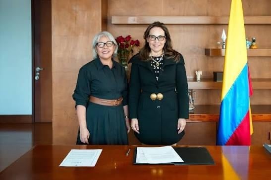 Con Viviane Morales al frente, los cristianos aterrizan con cargos de poder en el Ministerio de Educación Two women stand behind a desk with documents for signing, a Colombian flag visible to the right in a formal office setting. - Con Viviane Morales al frente, los cristianos aterrizan con cargos de poder en el Ministerio de Educación
