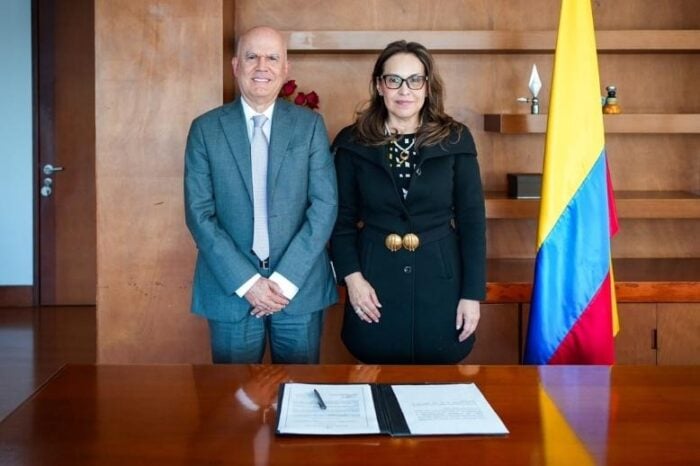 Con Viviane Morales al frente, los cristianos aterrizan con cargos de poder en el Ministerio de Educación Two formally dressed individuals stand beside a wooden desk with an open document, after signing a pact or agreement, beside a yellow-blue-red flag. - Con Viviane Morales al frente, los cristianos aterrizan con cargos de poder en el Ministerio de Educación