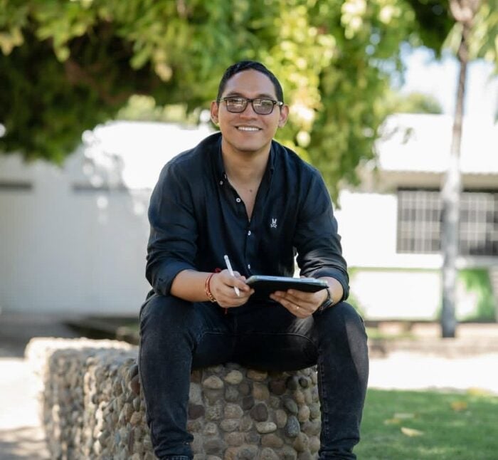 El capellán del Palacio tendrá nuevo rol: será la voz del presidente en 5 universidades públicas Smiling man wearing glasses and a dark button-down shirt sits on a stone bench outdoors, writing on a tablet with a stylus. - El capellán del Palacio tendrá nuevo rol: será la voz del presidente en 5 universidades públicas