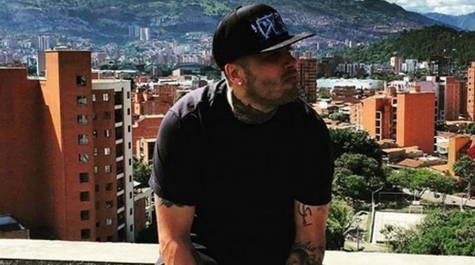 Man wearing a black T-shirt and cap sits on a rooftop, with a cityscape and distant mountains in the background. - La segunda oportunidad en Colombia que sacó a Nicky Jam de la pobreza y lo volvió un gigante del reguetón