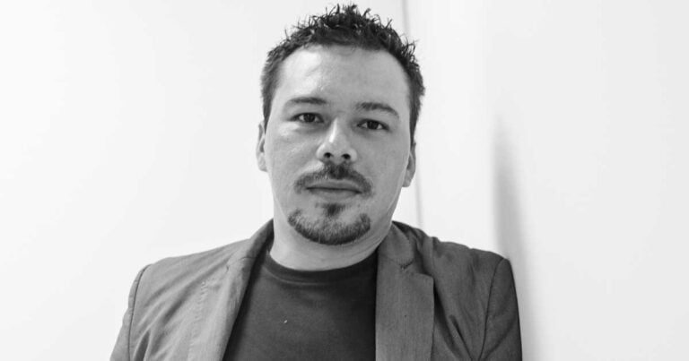 Portrait of a man with short dark hair and goatee, wearing a blazer over a dark shirt, leaning against a white wall. - La trampa de la batalla cultural