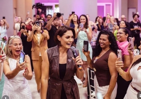 Cómo una atractiva mujer que venía de abajo de quien se enamoró Jair Bolsonaro se volvió el fenómeno político de Brasil A woman in a brown sleeveless coat speaks into a microphone as a cheering crowd gathers around her. - Cómo una atractiva mujer que venía de abajo de quien se enamoró Jair Bolsonaro se volvió el fenómeno político de Brasil
