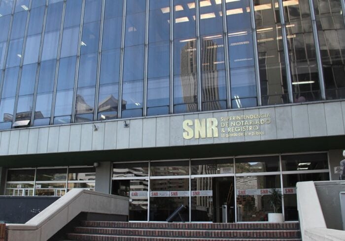Por orden del presidente Dela Espriella, Joaquín Méndez superintendente de Notariado Bloqueó entrega de notarías Front of a modern glass office building with the SNR sign above the entrance. - Por orden del presidente Dela Espriella, Joaquín Méndez superintendente de Notariado Bloqueó entrega de notarías