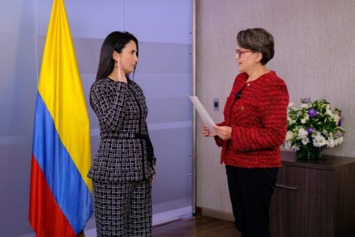 Dos abogadas del bufete De la Espriella Lawyers sin experiencia en el sector público saltan al alto gobierno Woman in a black-and-white suit takes an oath from a woman in a red jacket; Colombian flag in the background. - Dos abogadas del bufete De la Espriella Lawyers sin experiencia en el sector público saltan al alto gobierno