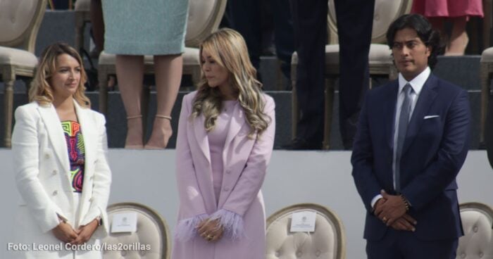 Three formally dressed people standing in a row on a stage outdoors, left and center women in light-colored outfits and a man in a dark suit on the right; chairs behind them suggest a ceremony setting. - La infidelidad de Nicolás Petro prendió el incendio que tiene enfrentadas y cerca de la cárcel a su esposa y a su ex