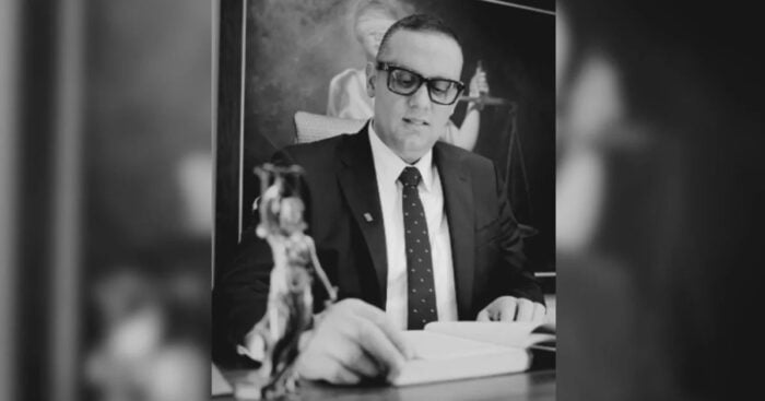 Black-and-white photo of a lawyer in a suit and glasses reading a document at his desk, with scales of justice in the background. - Derechos sin contrapesos: el punto ciego de la protección indígena.