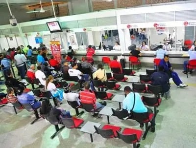 La ministra Elsa Noguera bloqueó el nuevo requisito para sacar licencias de conducción People seated in a crowded waiting area of a service center, facing rows of counters where staff assist customers. - La ministra Elsa Noguera bloqueó el nuevo requisito para sacar licencias de conducción