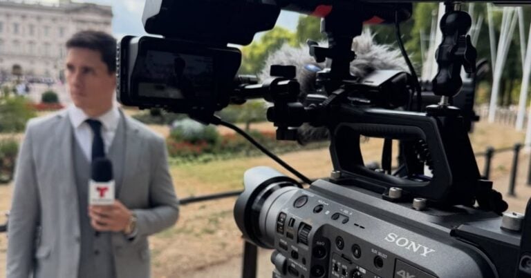 Las2orillas.co: Historias, voces y noticias de Colombia News reporter in a gray suit holding a microphone, filmed by a large professional camera outdoors. - Las2orillas.co: Historias, voces y noticias de Colombia