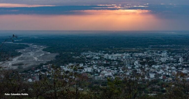 Las2orillas.co: Historias, voces y noticias de Colombia Sunset over a river valley with a city below, viewed from a hillside; orange light breaks through clouds. - Las2orillas.co: Historias, voces y noticias de Colombia