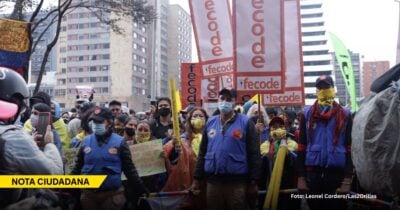 Crowd of protesters wearing blue vests and yellow masks, holding orange 'fecode' banners in a city street. A 'Nota Ciudadana' banner is visible in the foreground. - Las2orillas.co: Historias, voces y noticias de Colombia