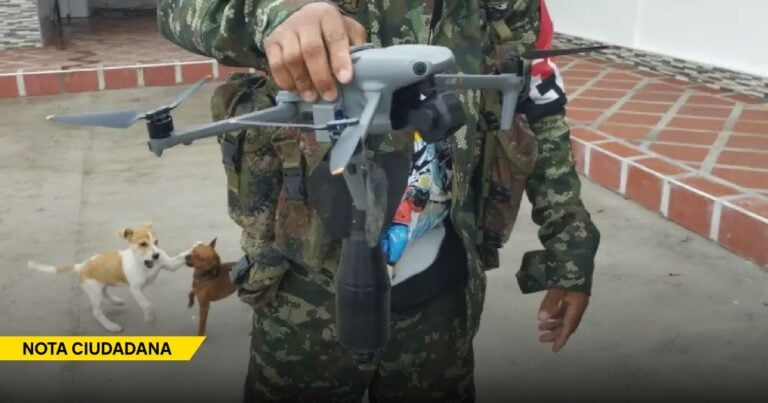 Person in camouflage uniform holding a gray quadcopter drone, with two small dogs barking in the background and a yellow 'NOTA CIUDADANA' banner at the bottom left. - Los drones artesanales que aterrorizan al Cauca y al Catatumbo sin que nadie pueda frenarlos
