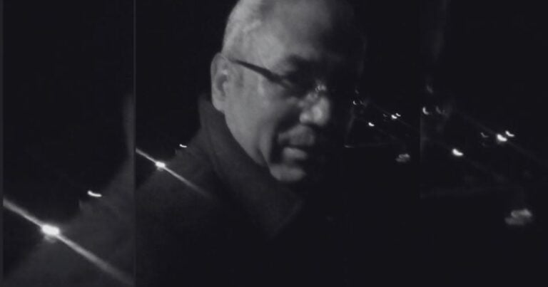 Las ciencias lúgubres revisitadas Man wearing glasses in a dark outdoor setting at night, with distant city lights in the background. - Las ciencias lúgubres revisitadas