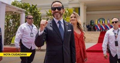 Man in a dark suit and sunglasses raises a clenched fist on a red carpet, with a smiling blonde woman in a red dress beside him and security nearby. - Las2orillas.co: Historias, voces y noticias de Colombia