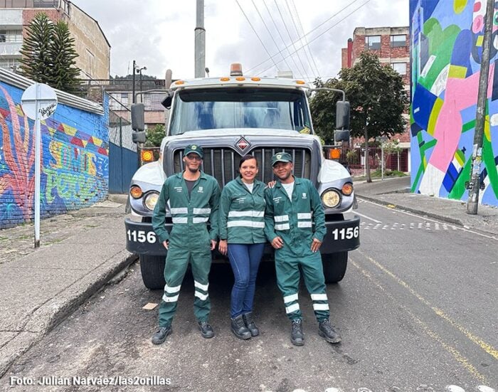 Three workers in green uniforms stand in front of a large truck on a graffiti-covered urban street. - Ella es Diana Barón, una de las tres mujeres que se abrieron camino al volante en el servicio de aseo de Bogotá