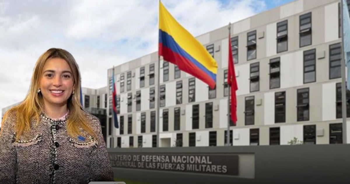 Smiling woman standing in front of a modern government building with the Colombian flag flying overhead. - Una mujer llegó al puesto clave para que las fuerzas militares puedan cumplir con sus misiones