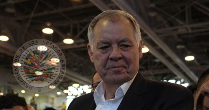 Older man in a suit at an event, with the circular seal of the Organization of American States nearby. - Las2orillas.co: Historias, voces y noticias de Colombia