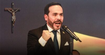 Las2orillas.co: Historias, voces y noticias de Colombia Man in a dark suit and blue tie speaks into a microphone with a clenched fist, a crucifix on the wall behind him. - Las2orillas.co: Historias, voces y noticias de Colombia