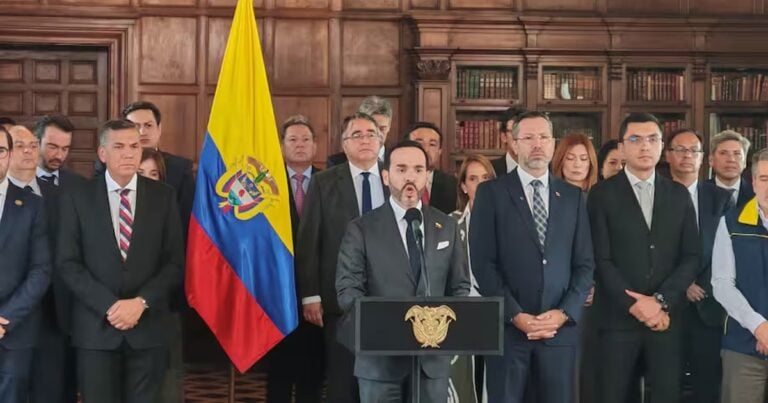 Las2orillas.co: Historias, voces y noticias de Colombia Group of formally dressed officials standing behind a podium with a gold eagle emblem, in a wood-paneled room, with a yellow-blue-red flag behind them, suggesting a formal government press event. - Las2orillas.co: Historias, voces y noticias de Colombia