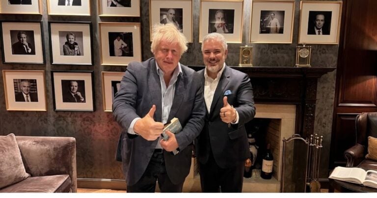 Las2orillas.co: Historias, voces y noticias de Colombia Two men in suits pose giving thumbs up in a stylish room with a wall of framed black-and-white portraits behind them. - Las2orillas.co: Historias, voces y noticias de Colombia