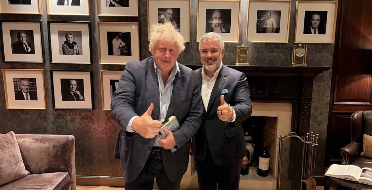 Two men in suits pose giving thumbs up in a stylish room with a wall of framed black-and-white portraits behind them. - El poder del ex presidente Iván Duque con su Fundación que trajo al ex ministro Boris Johnson a Colombia