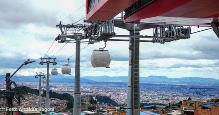 Dos familias ligadas al transporte bogotano junto a los bolivianos de Mi Teleférico operarán el cable de San Cristóbal Cable car gondolas travel along a hillside lift above Bogotá, with a dense cityscape and distant mountains under a cloudy sky. - Dos familias ligadas al transporte bogotano junto a los bolivianos de Mi Teleférico operarán el cable de San Cristóbal