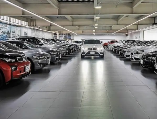 El encontrón entre Galan y los empresarios por el aumento del impuesto de Industria y Comercio White SUV drives toward the camera down the center aisle of a spacious car showroom with rows of parked cars on both sides. - El encontrón entre Galan y los empresarios por el aumento del impuesto de Industria y Comercio