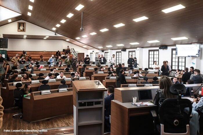 El encontrón entre Galan y los empresarios por el aumento del impuesto de Industria y Comercio Large legislative chamber with rows of wooden desks, microphones, and people conversing during a meeting or council session. - El encontrón entre Galan y los empresarios por el aumento del impuesto de Industria y Comercio