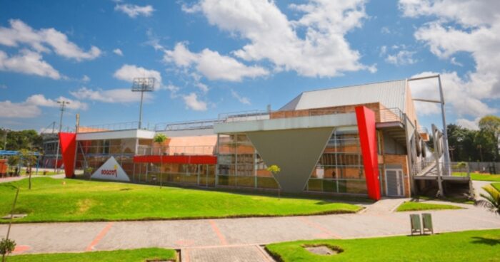 Después de diecisiete años y nueve contratos por más de .000 millones la Unidad Deportiva Salitre sigue igual Contemporary campus building with red angular supports, glass walls, and a grassy courtyard under a blue sky. - Después de diecisiete años y nueve contratos por más de .000 millones la Unidad Deportiva Salitre sigue igual