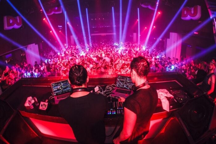 El encontrón entre Galan y los empresarios por el aumento del impuesto de Industria y Comercio Two DJs perform behind a mixing console at a crowded nightclub, with vibrant pink and blue laser lights sweeping over the dance floor. - El encontrón entre Galan y los empresarios por el aumento del impuesto de Industria y Comercio