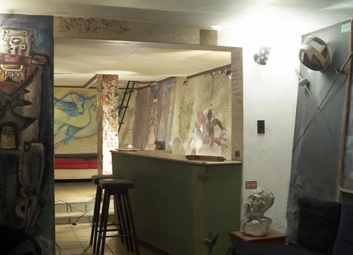 Fue un mexicano quien rescató de un sótano en Medellín los primeros murales que pintaron Botero y Obregón A small art-filled bar area with a green counter, two stools, and colorful mural walls in a basement-like room. - Fue un mexicano quien rescató de un sótano en Medellín los primeros murales que pintaron Botero y Obregón