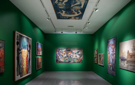 Fue un mexicano quien rescató de un sótano en Medellín los primeros murales que pintaron Botero y Obregón Green-walled art gallery with multiple framed paintings along the walls and a large artwork centered on the far wall under track lighting. - Fue un mexicano quien rescató de un sótano en Medellín los primeros murales que pintaron Botero y Obregón